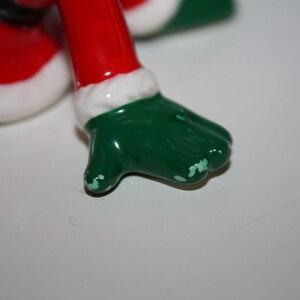 May include: A close-up of a ceramic Christmas figurine's foot. The foot is green with white paint chipping. The leg is red with a white trim. The figurine is likely a Santa Claus or elf.