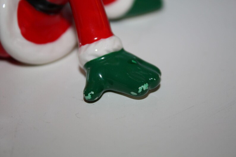 May include: A close-up of a ceramic Christmas figurine's foot. The foot is green with white paint chipping. The leg is red with a white trim. The figurine is likely a Santa Claus or elf.