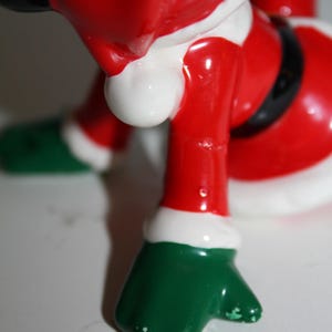 May include: A ceramic Santa Claus figurine, featuring a red suit with white trim and a black belt. The figurine has green gloves and boots. The image is a close-up of the lower body of the figurine.