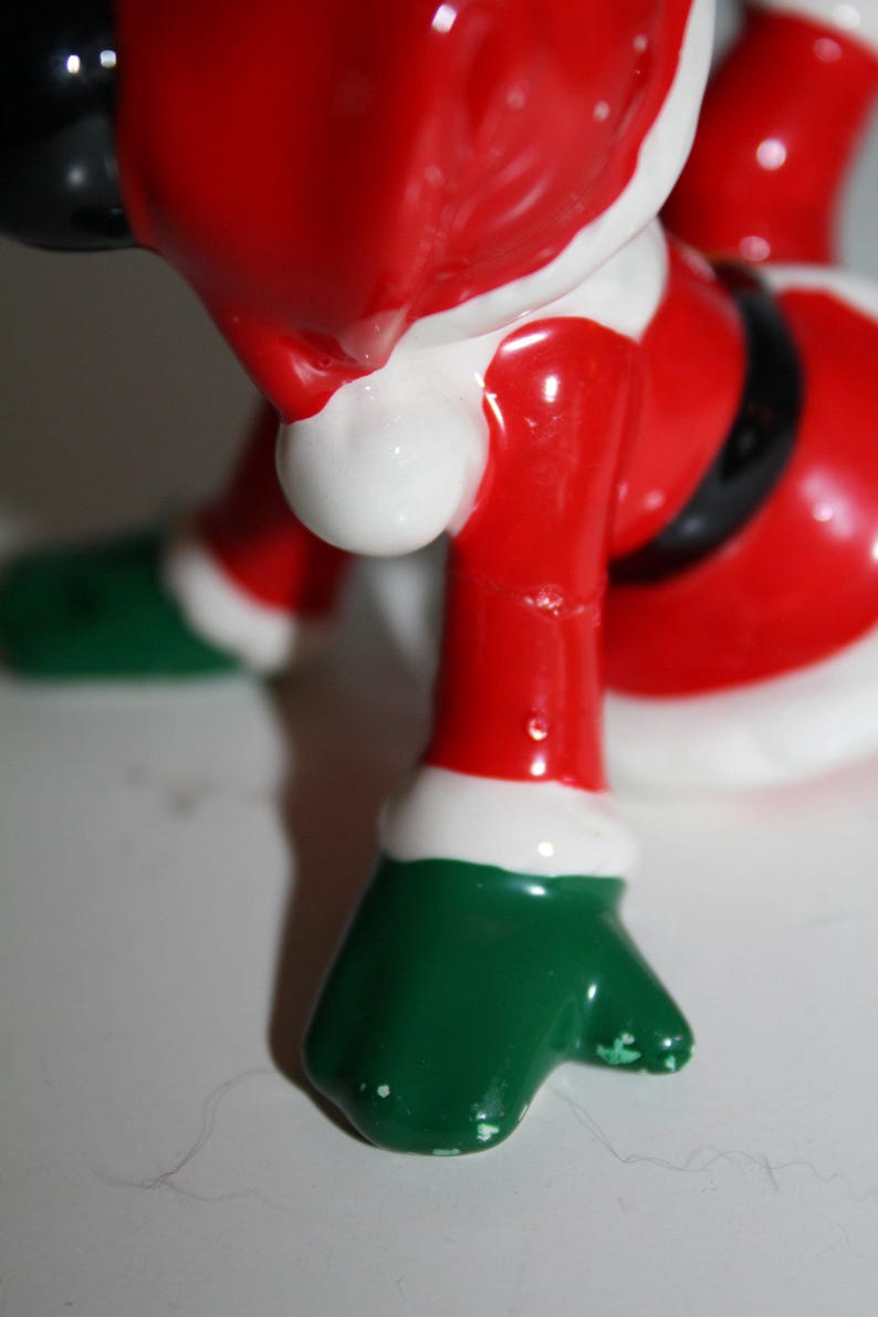 May include: A ceramic Santa Claus figurine, featuring a red suit with white trim and a black belt. The figurine has green gloves and boots. The image is a close-up of the lower body of the figurine.