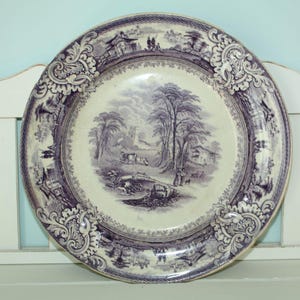May include: A round, antique decorative plate with a purple and white landscape scene. The plate has a detailed border with floral and architectural elements. The central image depicts trees, buildings, and figures, creating a vintage aesthetic.