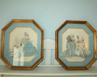 May include: Two framed prints featuring women in elegant gowns. The artwork is set within octagonal gold frames with a light blue matting. The women are depicted in shades of blue, white, and cream, with intricate details.