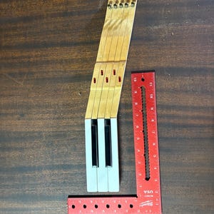 Puede incluir: Un conjunto de tres teclas de piano con tallos de madera clara y cubiertas blancas, cada una con una tecla negra. Una escuadra de metal rojo en forma de L está colocada al lado, mostrando una longitud total de unos 30,5 cm.