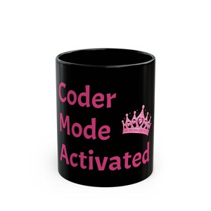 May include: A black ceramic mug featuring the pink text Coder Mode Activated stacked in three lines on the front. To the right of the text is a pink graphic illustration of a crown with small heart shapes and star accents integrated into the design.