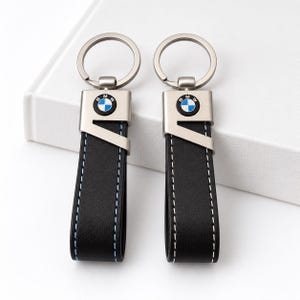 Könnte beinhalten: Zwei schwarze Schlüsselanhänger aus Leder mit einem silberfarbenen Metallaufsatz und dem BMW-Logo. Ein Anhänger hat blaue Nähte, der andere weiße Nähte. Beide verfügen über einen runden Metallring zur Befestigung von Schlüsseln.