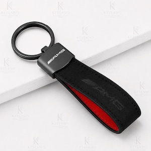 Puede incluir: Un llavero negro con un anillo metálico circular unido a un conector metálico rectangular con el logotipo AMG en blanco. La correa es de material negro con un forro interior rojo y el logotipo AMG grabado en la superficie.