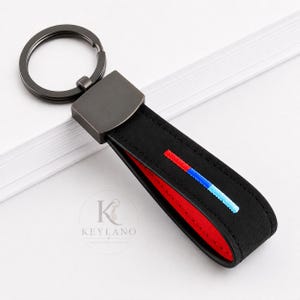 Peut inclure: Un porte-clés noir avec un anneau en métal foncé et un connecteur rectangulaire attaché à une sangle noire à doublure rouge. La sangle présente un détail brodé horizontal en rouge, bleu foncé et bleu clair. Le texte KEYLANO est visible.