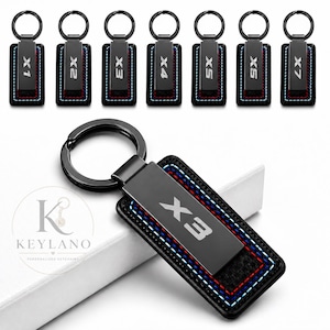 May include: A rectangular black metal keychain featuring a central plate engraved with model numbers X1, X2, X3, X4, X5, or X7. The plate is bordered by black material with decorative red, white, and blue stitching and a black metal ring.