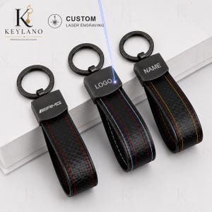 Könnte beinhalten: Drei schwarze Schlüsselanhänger mit Carbonfaser-Struktur, Metallringen und rechteckigen Beschlägen. Der erste zeigt das AMG-Logo, der zweite eine Lasergravur des Wortes LOGO und der dritte das Wort NAME, das in die Metalloberfläche eingraviert ist.