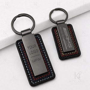 May include: Two rectangular keychains with black metal hardware and black leather-like borders featuring multi-coloured stitching. One keychain displays the text YOUR LOGO CUSTOM, while the other features the Mercedes-Benz AMG logo and text.