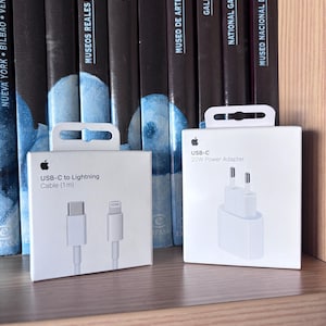 Könnte beinhalten: Zwei weiße Apple-Produktverpackungen. Die linke Schachtel zeigt ein USB-C auf Lightning Kabel (1m) mit einer Abbildung der Kabelenden. Die rechte Schachtel zeigt ein 20W USB-C Netzteil mit einer Abbildung des weißen Wandsteckers.
