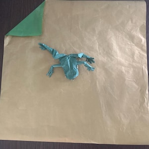 May include: A green origami frog crafted from paper, positioned in the center of a large, square sheet of tan-colored paper that features a single folded corner revealing a bright green underside, creating a contrast in color and texture.