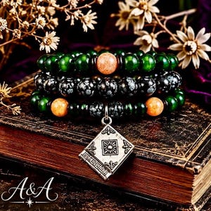 May include: A multi-strand beaded bracelet featuring polished dark green beads, black snowflake obsidian beads, and light orange sunstone beads. A silver-toned metal charm shaped like a square book with an embossed sun design hangs from the center.