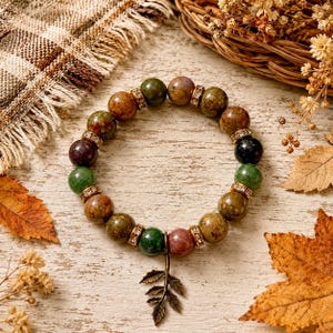 May include: A beaded bracelet featuring round, multi-colored stone beads in shades of green, brown, and black. The bracelet is accented with small, gold-toned metal spacers embedded with clear crystals and a dangling bronze-toned metal leaf charm.