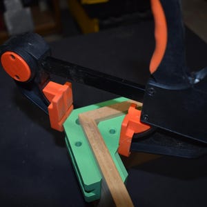 May include: A right-angle corner clamp tool featuring a bright green L-shaped base and orange pressure pads. The tool is constructed with black plastic components and a metal bar, securely holding two pieces of light-colored wood together at a ninety-degree angle.