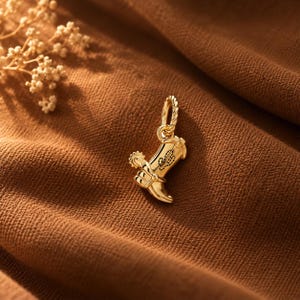 May include: A gold-toned metal charm shaped like a cowboy boot, featuring a small spur on the heel and a delicate stitched pattern detail on the side. The charm includes a small circular jump ring at the top for attachment to a chain or bracelet.