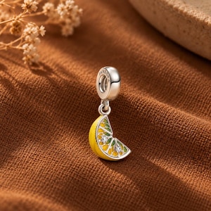 Può includere: Un charm in argento a forma di spicchio di agrume con dettagli in smalto giallo e verde e due piccole pietre trasparenti incastonate negli spicchi del frutto. L'anello superiore è inciso con la parola PANDORA e ha una finitura metallica lucida.