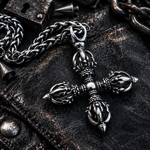 May include: A silver-toned metal pendant shaped like a double vajra, featuring intricate, symmetrical prongs and a central sphere. The pendant is attached to a thick, woven silver-toned metal chain, showcasing a polished finish with dark oxidation.