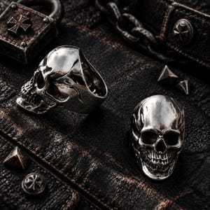 May include: Two polished silver-toned rings shaped like human skulls. One ring is positioned in a side profile view, while the other is displayed from a front-facing perspective, showcasing detailed eye sockets, nasal cavities, and teeth.