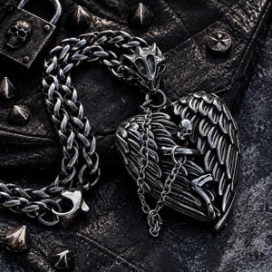 Può includere: Una collana in metallo color argento con una spessa catena intrecciata e un grande ciondolo a forma di cuore. Il ciondolo è dettagliato con ali scolpite e un piccolo teschio. Una catena secondaria più sottile collega il ciondolo alla catena principale.