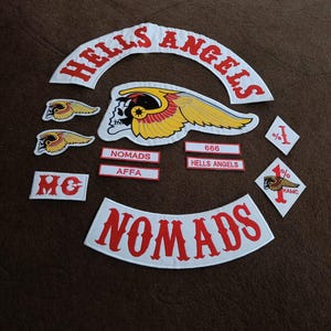 May include: A collection of embroidered patches featuring the text HELLS ANGELS and NOMADS in red on white fabric. Includes a central winged skull emblem, smaller winged skull patches, rectangular name tags, and diamond-shaped patches with 1% and HAMC text.