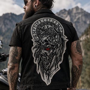 May include: A black denim vest featuring a large, detailed back patch with a graphic illustration of a bearded figure wearing a helmet, accented by a single red eye and surrounded by circular runic text in a white and black color scheme.