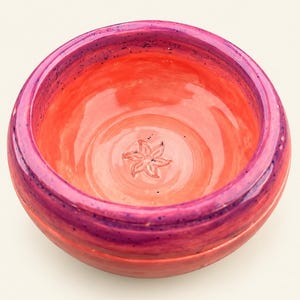 Könnte beinhalten: Eine runde Keramikschale mit einem leuchtend korallenorangen Inneren und einem tiefvioletten Rand. In der Mitte der Schale befindet sich ein kleines, geprägtes Blumenmotiv mit sechs Blütenblättern. Die Außenseite verläuft von einem violetten oberen Teil zu einer korallenorangen Basis.