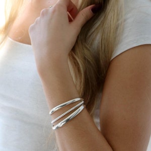 May include: Silver cuff bracelets. The image shows three silver cuff bracelets on an arm. The top and bottom cuffs are smooth, while the middle cuff has a row of small, sparkling stones. The bracelets are worn on the left wrist.