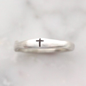 May include: A silver ring with a small, dark cross etched into the band. The ring has a slightly textured surface and a simple, elegant design. The cross is centered on the ring.