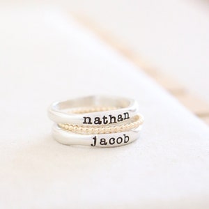 May include: A stack of three rings. The top and bottom rings are silver with the names "nathan" and "jacob" stamped on them. A gold beaded ring sits between them. The rings are on a white surface.