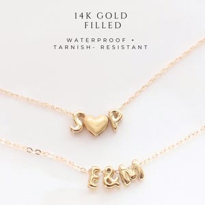 May include: Two gold necklaces with initial charms. The top necklace features the letters "S" and "P" with a heart charm in between. The bottom necklace has the letters "E", "&", and "M". The necklaces are 14k gold filled, waterproof, and tarnish-resistant.