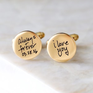 May include: A pair of gold-colored cufflinks with handwritten inscriptions. One cufflink reads "Always forever 03.28.26", and the other says "i love you" with a heart symbol. The cufflinks are round and have a shiny finish.