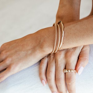 May include: A close-up of a hand wearing three gold bangles and a gold beaded ring. The hand is resting on a white surface.