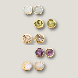 May include: Five pairs of stud earrings featuring raw, uncut gemstones set in gold-toned metal. The collection includes white, light green, clear, purple, and iridescent stones, each with an irregular, organic shape and a smooth, polished gold border.