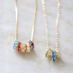 May include: Two gold necklaces featuring small, circular charms. The left necklace has five charms with light blue, red, peach, white, and purple stones. The right necklace has three charms with light green, blue, and clear stones.
