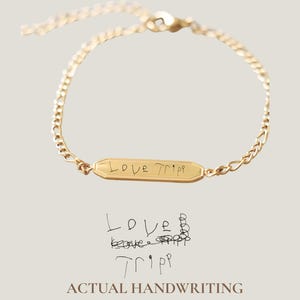 May include: A gold-colored bracelet with a chain design and a rectangular pendant. The pendant is engraved with the words "Love Tripp" in a handwritten style. The image also includes the text "Actual Handwriting".