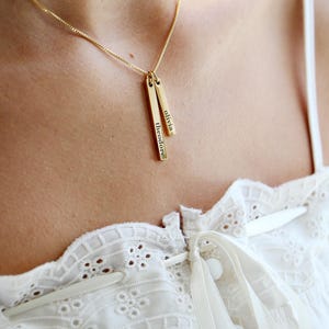 May include: A gold-toned necklace featuring two vertical bar pendants suspended from a delicate chain. Each rectangular bar is engraved with a name in a black sans-serif font, with one bar reading the name theodore and the other reading olivia.