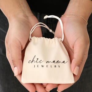 以下が含まれることがあります： 「chic mama JEWELRY」の文字が黒い筆記体でプリントされた、クリーム色の小さな巾着袋。 2つの手で開かれており、手首には繊細なゴールドのブレスレットが見えます。