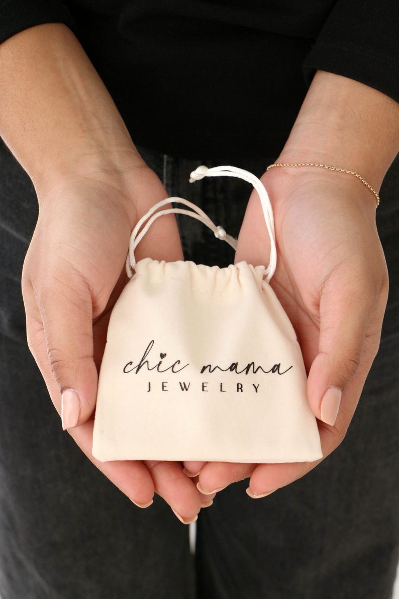 以下が含まれることがあります： 「chic mama JEWELRY」の文字が黒い筆記体でプリントされた、クリーム色の小さな巾着袋。 2つの手で開かれており、手首には繊細なゴールドのブレスレットが見えます。