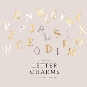 Op de afbeelding: Een verzameling letterbedels van goud- en zilverkleurig metaal met diverse letters van het alfabet. De tekst luidt: SILVER + GOLD LETTER CHARMS A - Z (NO U OR Y). Elke bedel is voorzien van een klein oogje aan de bovenzijde.