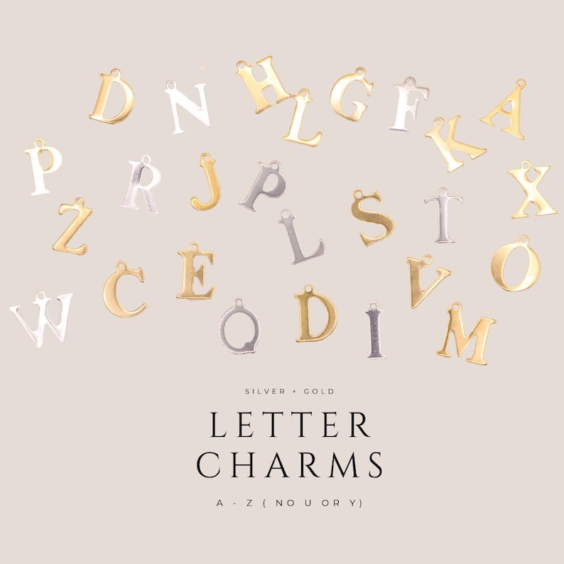 Op de afbeelding: Een verzameling letterbedels van goud- en zilverkleurig metaal met diverse letters van het alfabet. De tekst luidt: SILVER + GOLD LETTER CHARMS A - Z (NO U OR Y). Elke bedel is voorzien van een klein oogje aan de bovenzijde.
