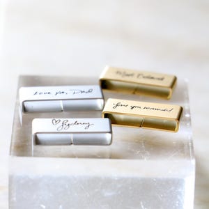 Może przedstawiać: Cztery prostokątne, spersonalizowane pendrive'y z odręcznymi wiadomościami. Dwa są srebrne, a dwa złote. Wiadomości to "love ya, Dad", "Sydney", "love you so much!" i "To Joel, Beloved". Pendrive'y są wyświetlane na przezroczystym, prostokątnym bloku.