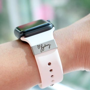 Może przedstawiać: Czarny smartwatch z białym paskiem i jasnor&oacute;żowym paskiem. Pasek ma wygrawerowane imię "Sydney" na srebrnym metalowym elemencie. Zegarek jest noszony na nadgarstku.
