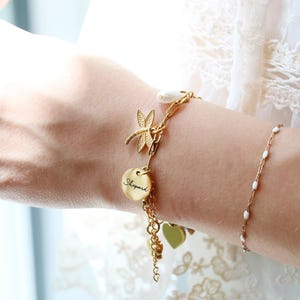 Op de afbeelding: Een goudkleurige bedelarmband met een libellebedel, een ronde schijf gegraveerd met de naam Shepard en een wit parelaccent. De armband is opgebouwd uit een mix van kettingstijlen en bevat extra hartvormige bedels.
