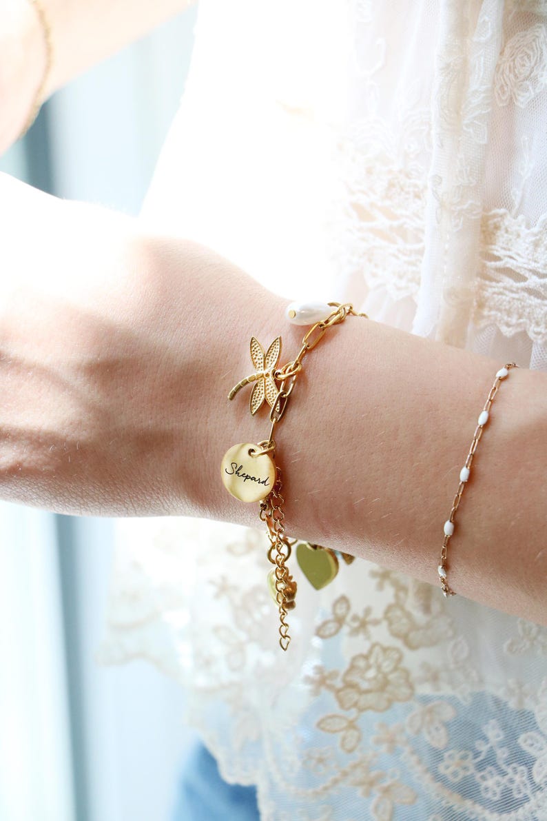 Op de afbeelding: Een goudkleurige bedelarmband met een libellebedel, een ronde schijf gegraveerd met de naam Shepard en een wit parelaccent. De armband is opgebouwd uit een mix van kettingstijlen en bevat extra hartvormige bedels.