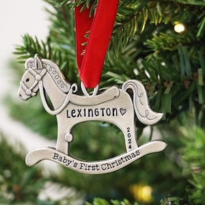 以下が含まれることがあります： 赤いリボンが付いた、銀色のロッキングホースのクリスマスオーナメント。「Lexington」と「Baby's First Christmas 2024」という名前が刻まれています。緑のクリスマスツリーに飾られています。