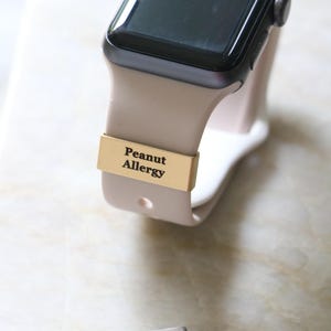 Może przedstawiać: Smartwatch z beżowym paskiem i czarnym ekranem. Złoty pasek z napisem "Peanut Allergy" jest przymocowany do paska zegarka. Osobna srebrna etykieta głosi "John Smith Typ 1 Diabetyk."