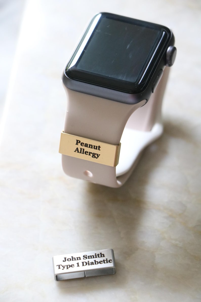 Może przedstawiać: Smartwatch z beżowym paskiem i czarnym ekranem. Złoty pasek z napisem "Peanut Allergy" jest przymocowany do paska zegarka. Osobna srebrna etykieta głosi "John Smith Typ 1 Diabetyk."