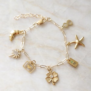 Op de afbeelding: Een gouden schakelarmband met bedels in de vorm van een halve maan, een palmboom, een koffer met een hartje, een klavertjevier met een steentje, een rechthoekig plaatje met de letter M en een groene steen, een zeester en een oneindigheidssymbool.