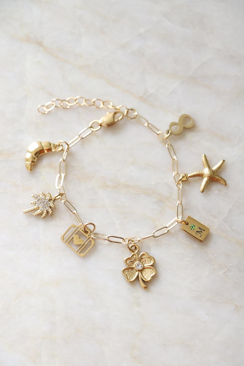Op de afbeelding: Een gouden schakelarmband met bedels in de vorm van een halve maan, een palmboom, een koffer met een hartje, een klavertjevier met een steentje, een rechthoekig plaatje met de letter M en een groene steen, een zeester en een oneindigheidssymbool.
